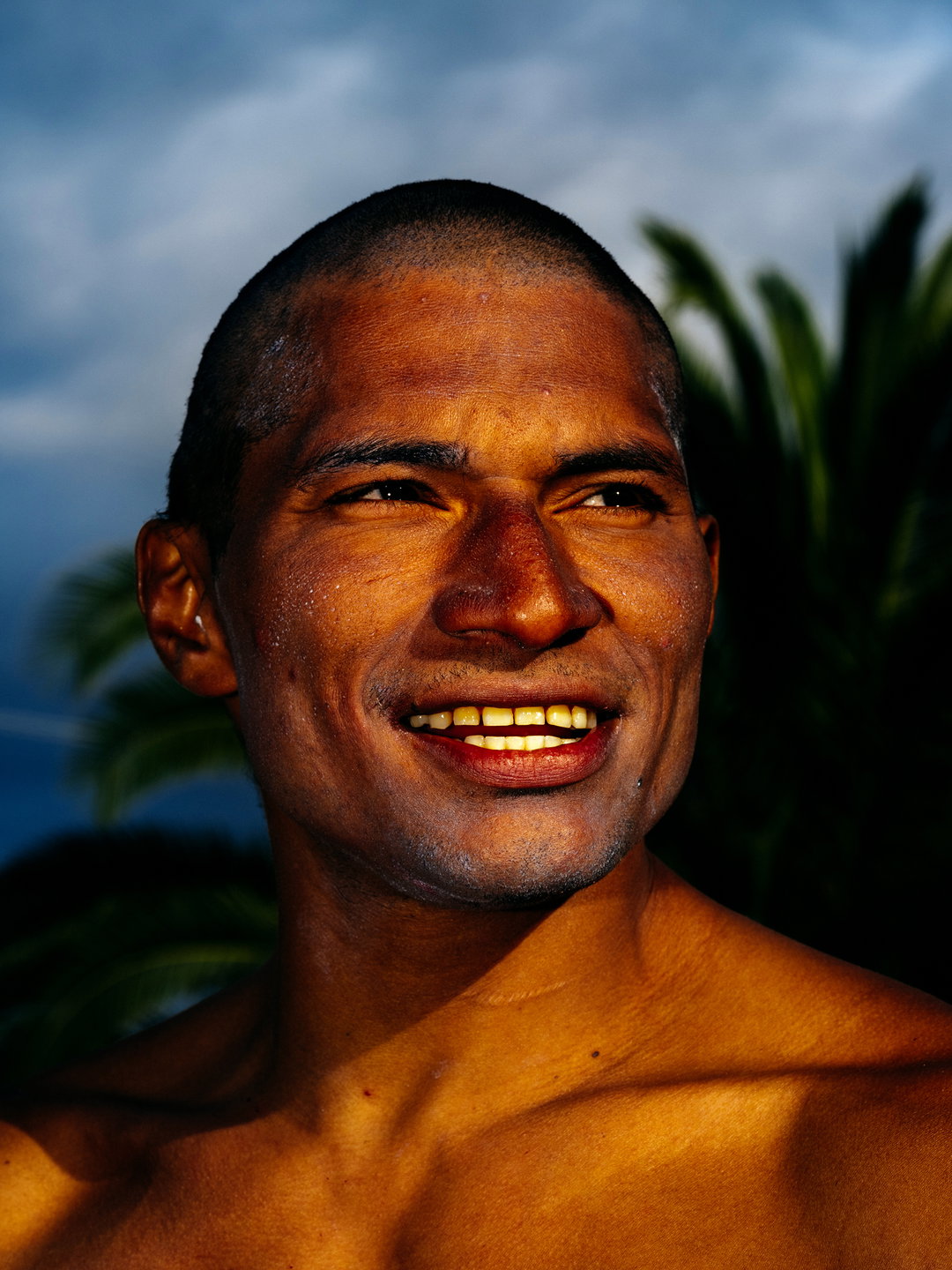 A close-up portrait of a person smiling and looking off-camera. They are lit by warm, golden light against a backdrop of a blue sky and dark green palm fronds.