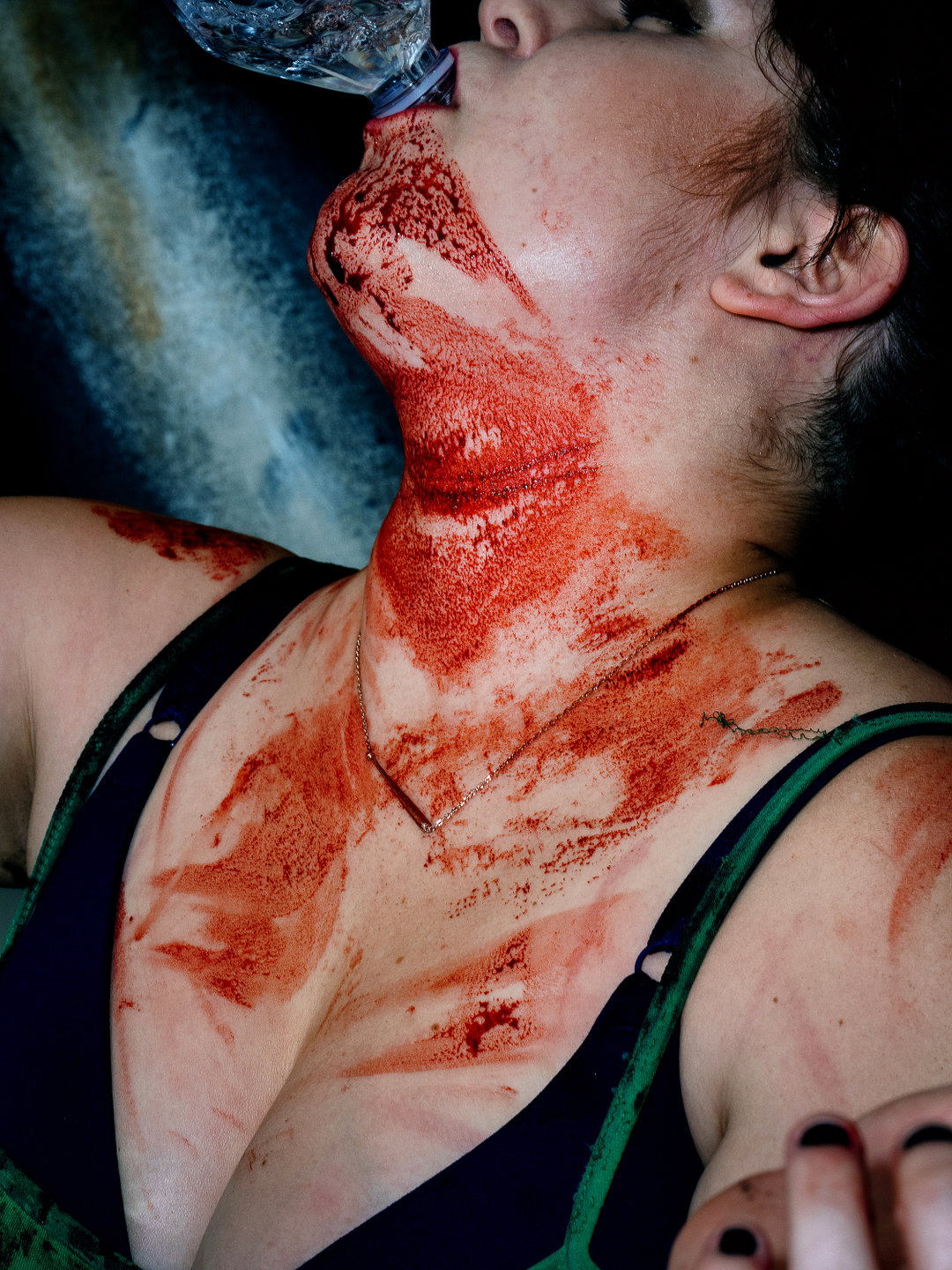 A person drinks from a plastic water bottle, their chin, neck and chest covered in streaks of red makeup. They wear a dark top with green straps and a thin gold chain necklace.