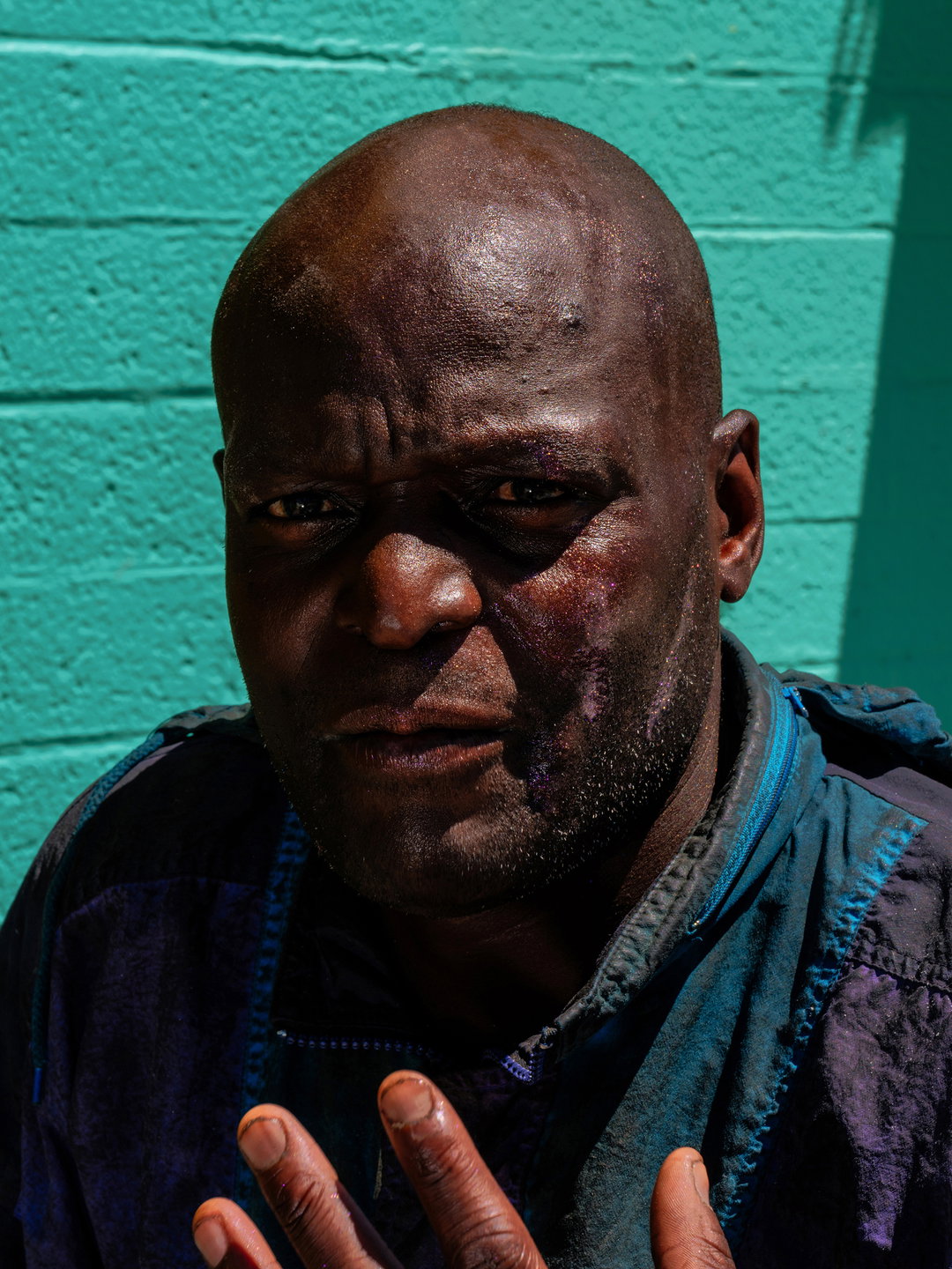 A close-up portrait of a person looking directly at the camera. They wear a dark blue jacket and stand against a bright turquoise brick wall.