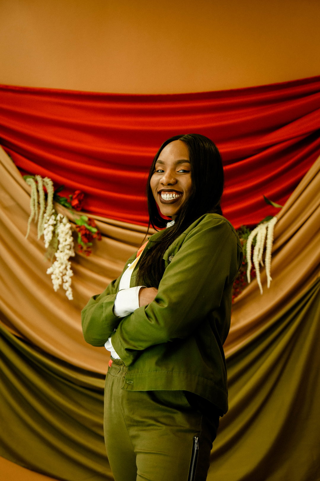 A person smiles at the camera with their arms folded, wearing an olive green utility suit over a white shirt. They stand before a backdrop of draped red, ochre and green fabric.