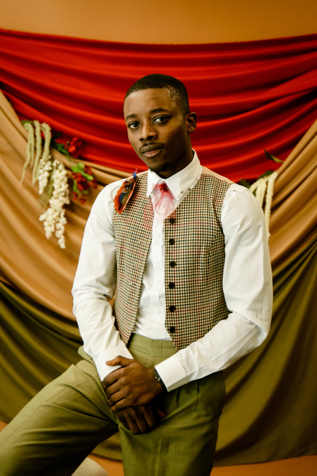 A person looks directly at the camera, wearing a white shirt, a checked waistcoat and olive green trousers. They sit against a backdrop of draped red, gold and green fabric.