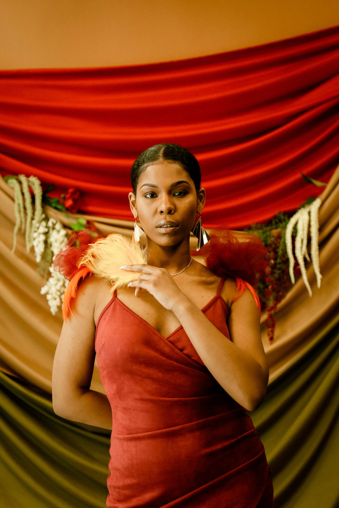 A person in a red velvet dress looks directly at the camera, holding a yellow feather to their chin. They stand before a backdrop of draped red, gold and green fabric adorned with white flowers.