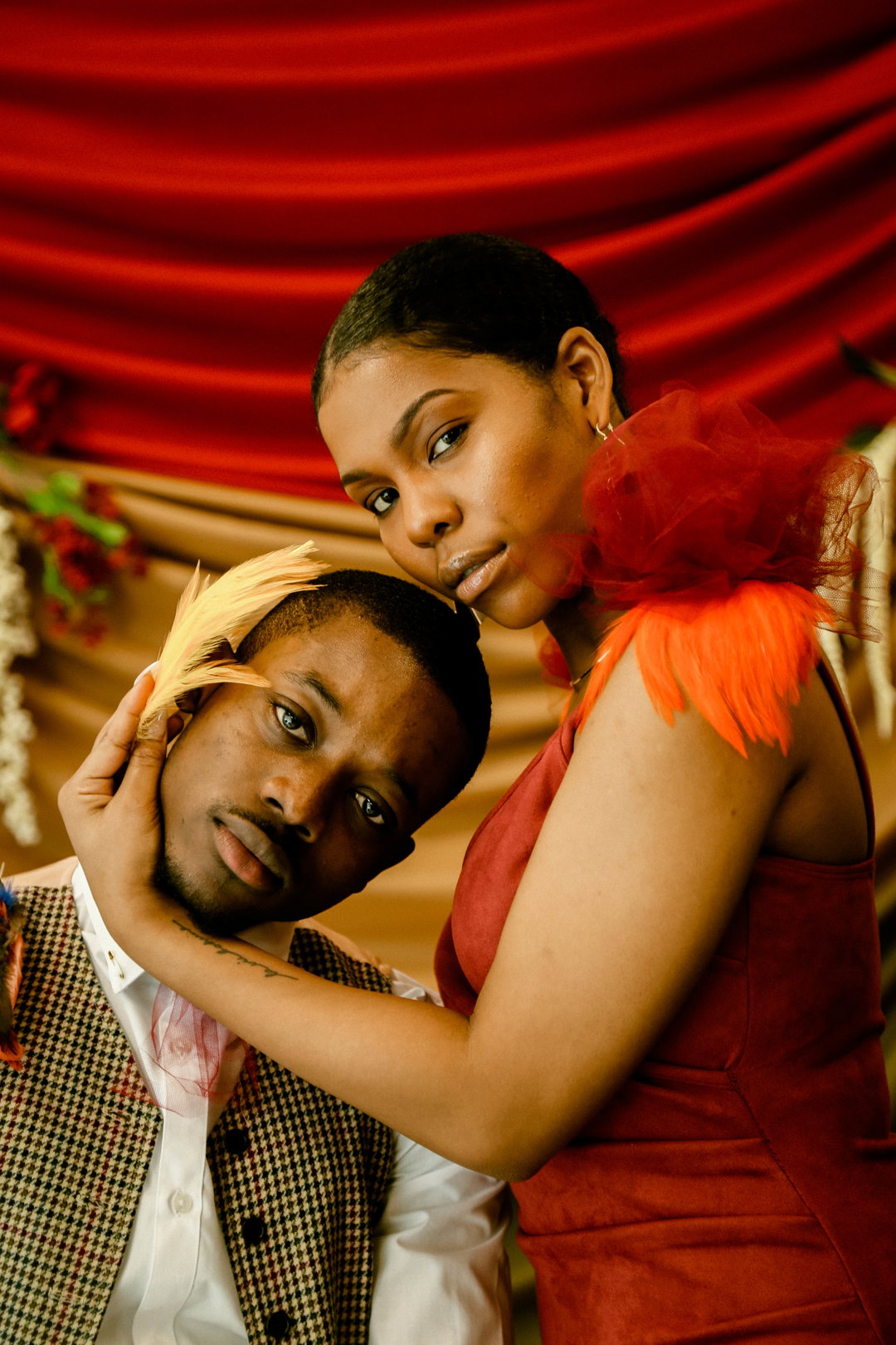 Two people pose closely together against a backdrop of draped red and gold fabric. One person wears a red dress with a voluminous orange tulle detail on the shoulder, while the other wears a checked waistcoat and a yellow feather in their hair.
