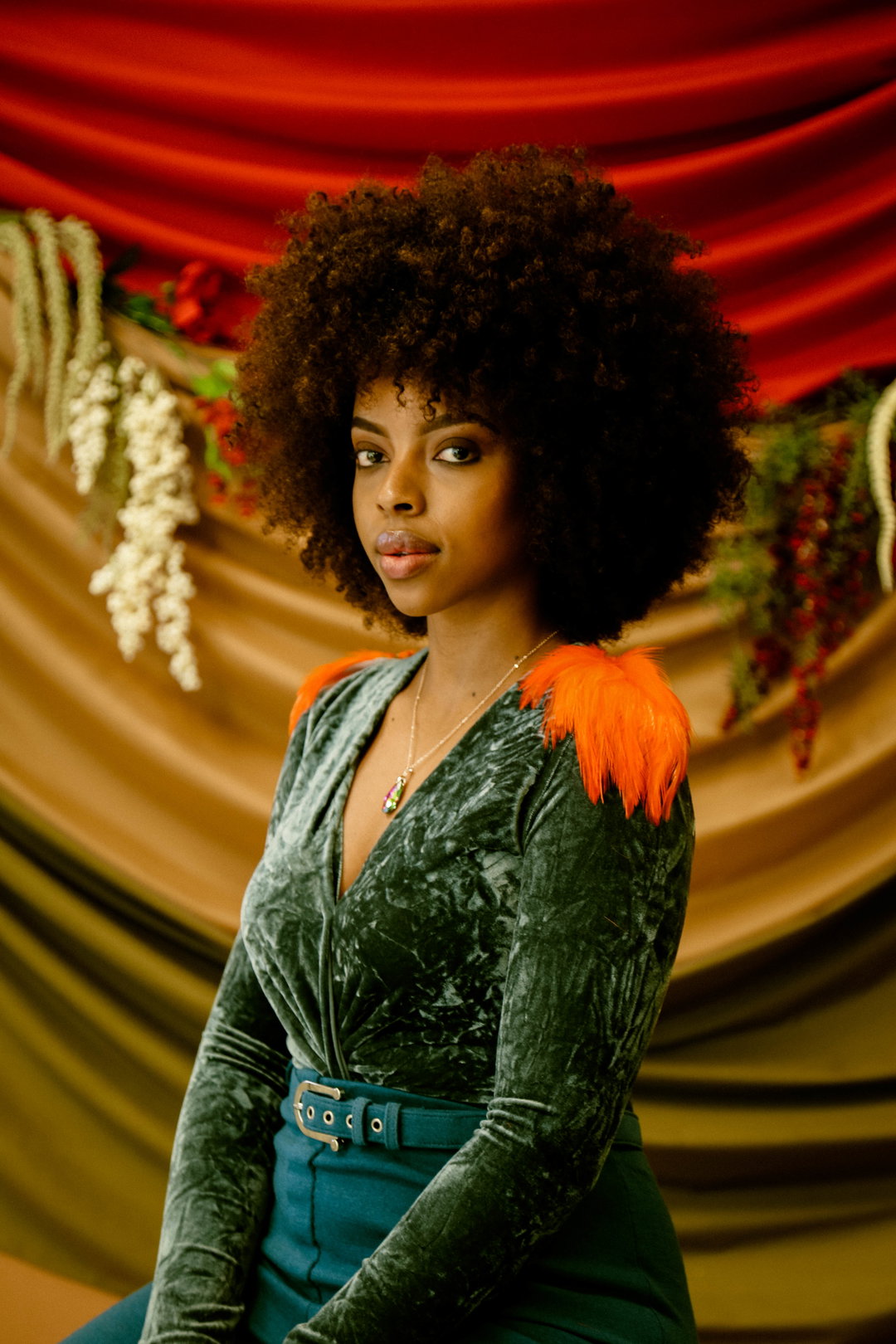 A person with a large afro looks directly at the camera. They wear a green velvet top with bright orange faux-fur shoulder details and a matching green belt.