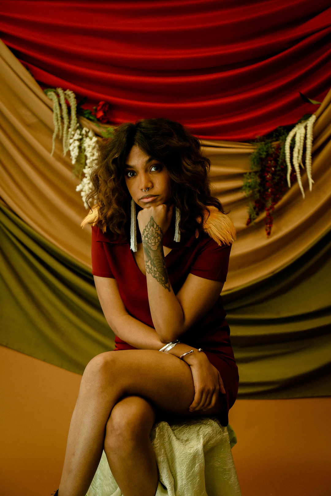 A person with curly hair and a tattoo on their forearm sits with their chin resting on their hand. They wear a burgundy dress and long, shimmering earrings against a backdrop of draped red, gold and green fabric.
