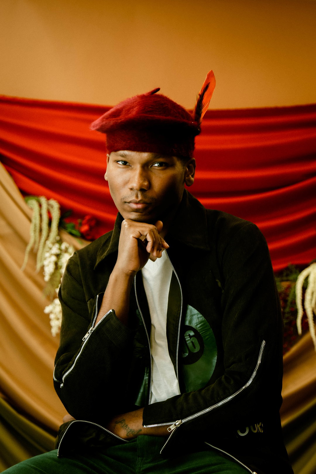 A person looks directly at the camera, resting their chin on their hand. They wear a black jacket over a white T-shirt and a red beret with a single orange feather.