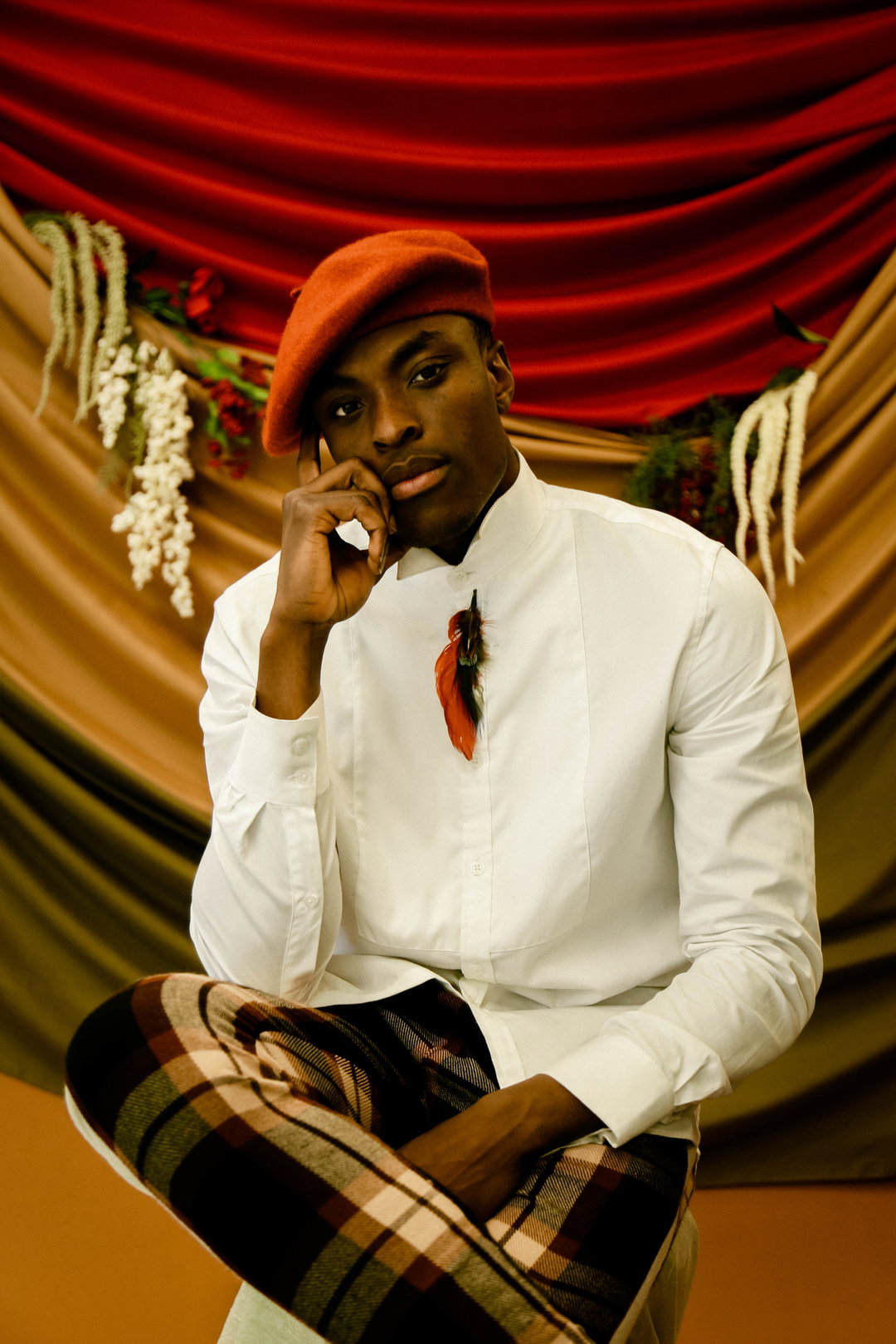 A person wears a red beret, a white button-down shirt and plaid trousers. They lean their chin on their hand, posing against a backdrop of draped red and gold fabric adorned with white flowers.