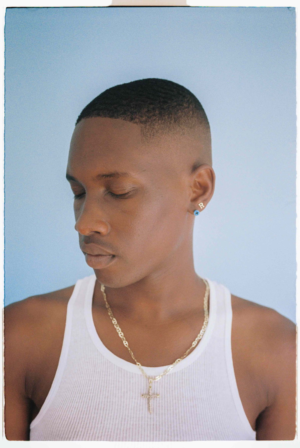 A person with a short fade haircut looks down and to the side against a plain blue background. They wear a white ribbed vest and a gold chain with a cross pendant.
