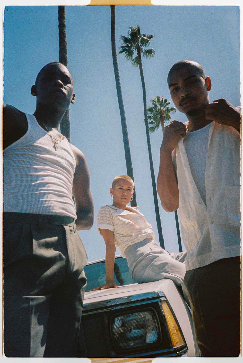 Three people pose together outdoors against a clear blue sky and tall palm trees. Two people stand in the foreground wearing white tops, while a third person with short cropped hair leans on the bonnet of a white car.